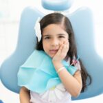 Child sitting in a dental chair with a hand on their cheek, suggesting tooth sensitivity.