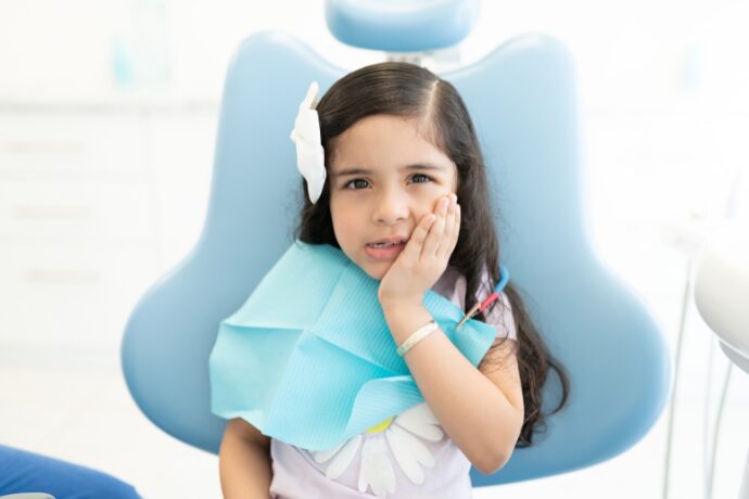 Child sitting in a dental chair with a hand on their cheek, suggesting tooth sensitivity.