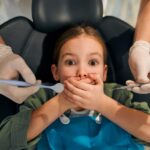 A young child covering her mouth in a dental chair while a pediatric dentist examines her teeth, representing sudden tooth pain and dental anxiety in cold weather.
