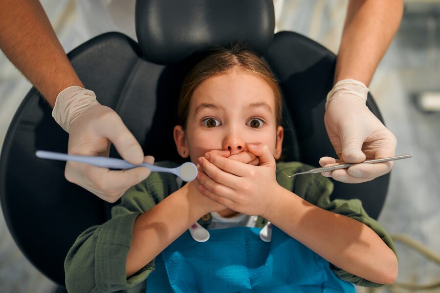 A young child covering her mouth in a dental chair while a pediatric dentist examines her teeth, representing sudden tooth pain and dental anxiety in cold weather.