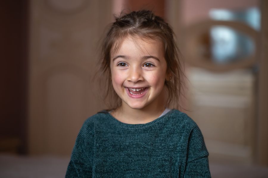 Young girl smiling at camera wearing teal shirt in indoor hallway setting.