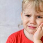 Young child in red shirt touching cheek, appearing to have tooth pain or discomfort.