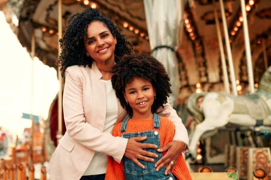 Woman and child smiling at carnival with carousel horses in warm, festive lighting background.