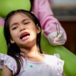 Young girl receiving dental examination in pediatric dentist's chair with dentist in pink attire.