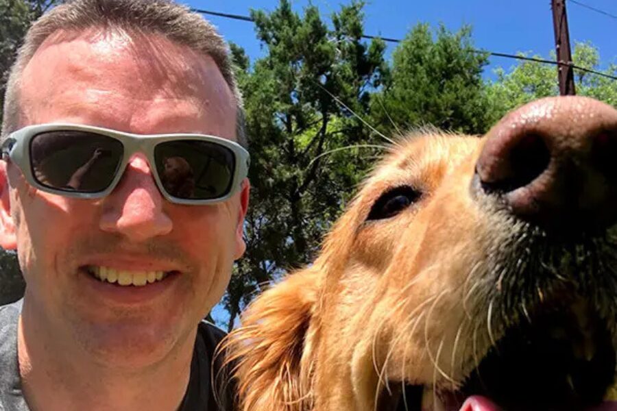 Man wearing sunglasses smiling next to a goat outdoors in sunny setting.