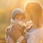 Mother holding toddler outdoors during golden hour sunset, both warmly dressed in winter clothing.