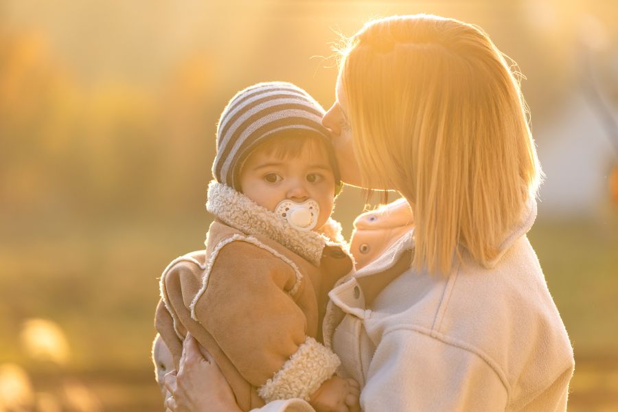 Mother holding toddler outdoors during golden hour sunset, both warmly dressed in winter clothing.