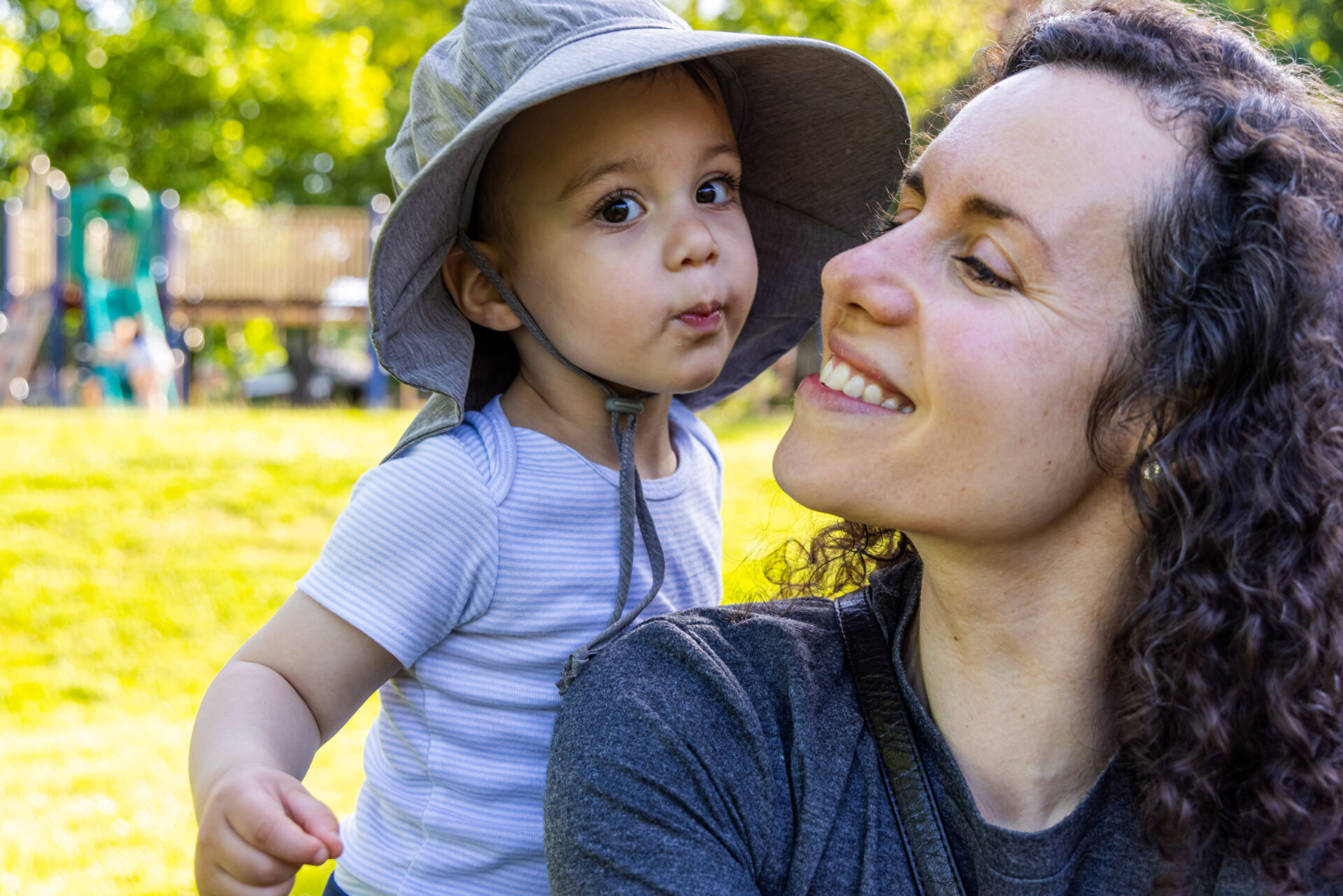 Mother holding toddler wearing sun hat in sunny park setting.